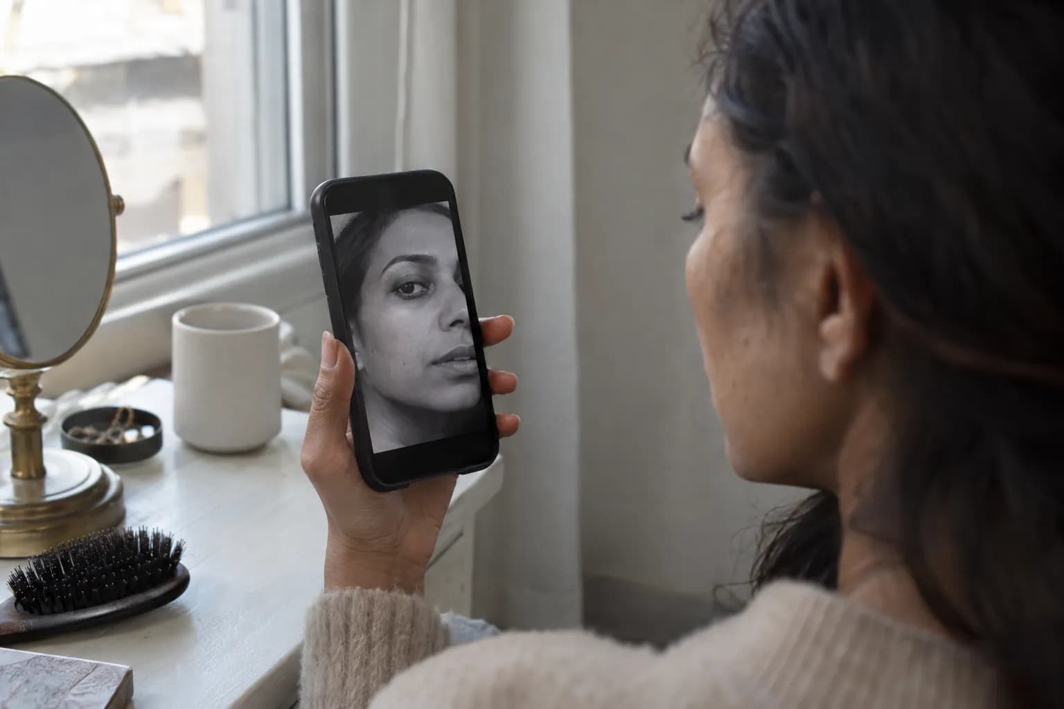 Woman beside a window checking a grayscale selfie to compare the depth of her skin, hair and eyes.
