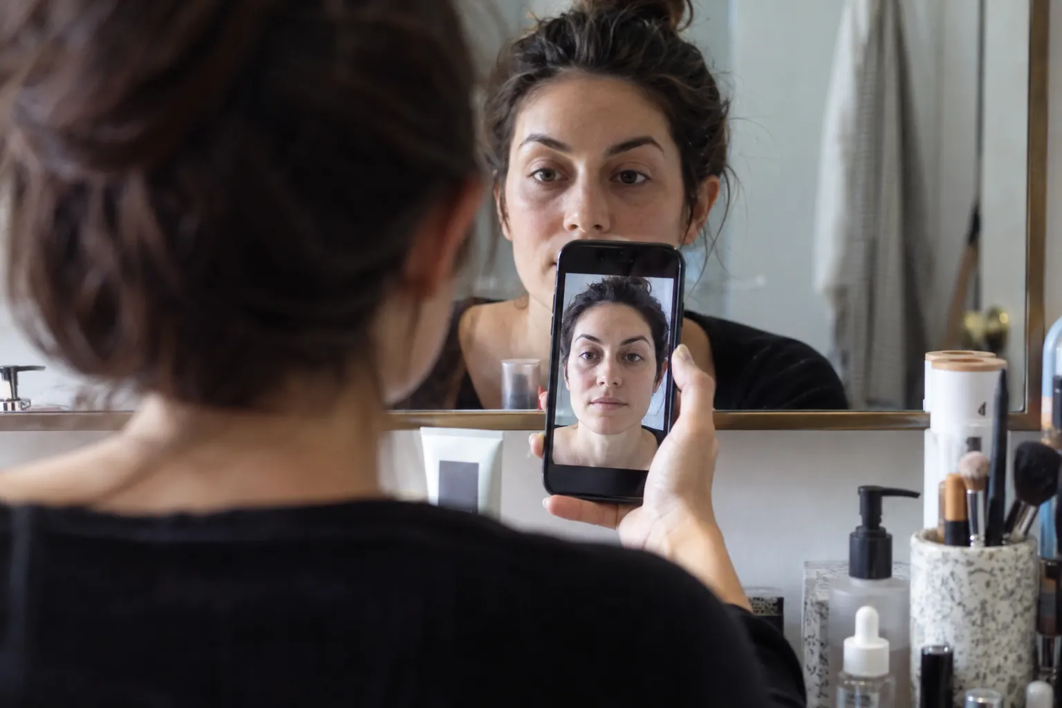 Woman checking her bare eyelids in an eye-level mirror beside a front-facing photo on her phone.