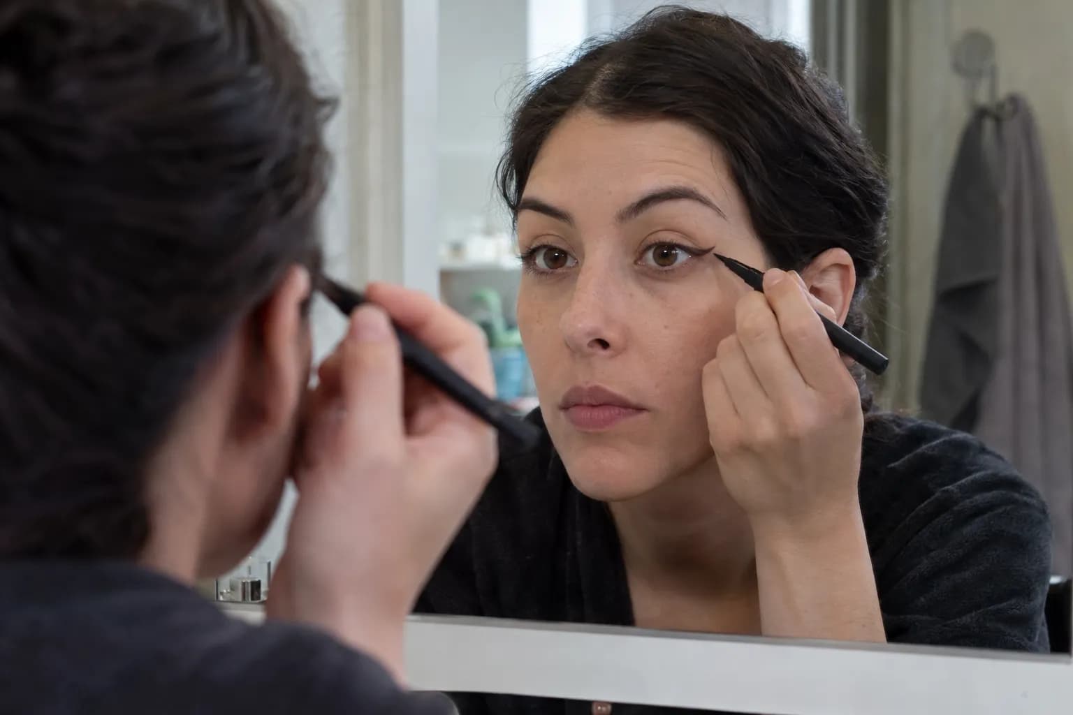 Woman with hooded eyes mapping a fine eyeliner wing in a mirror with her eyes open and brows relaxed.