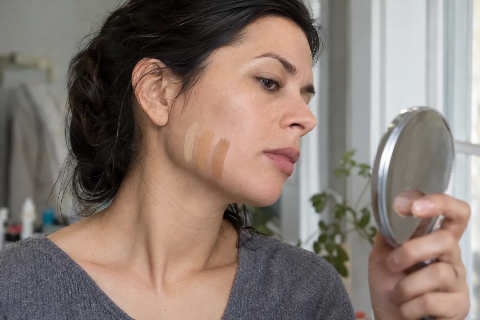 Woman examining three foundation swatches across her jaw beside a window in indirect daylight.