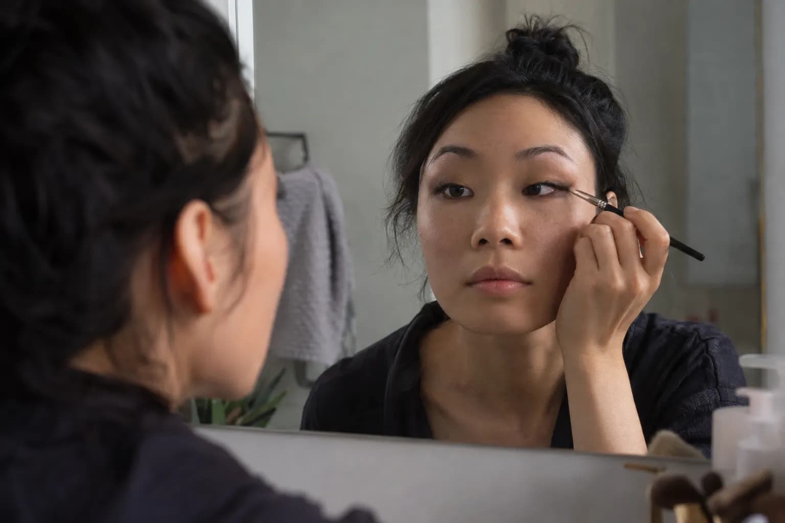 Woman with monolid eyes applying outer-corner eyeliner over warm taupe shadow while looking into a mirror.