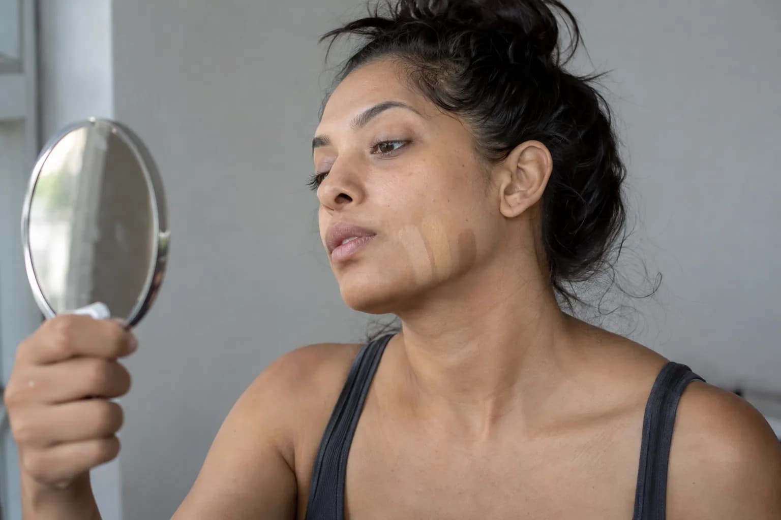 Woman with tan skin comparing three foundation swatches along her jaw beside a window.